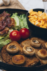 Close shot of tasty and fresh traditional meal with toast, scrambled eggs, fried bacon, mushrooms, green salad and cherry tomatoes.