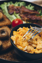 Close shot of tasty and fresh traditional meal with toast, scrambled eggs, fried bacon, mushrooms, green salad and cherry tomatoes.