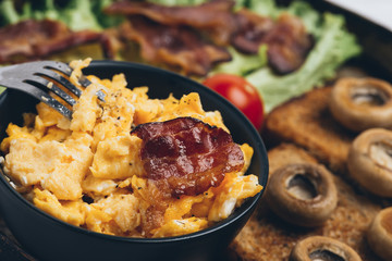 Close shot of tasty and fresh traditional meal with toast, scrambled eggs, fried bacon, mushrooms, green salad and cherry tomatoes.
