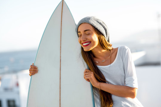Portrait of a young smiling woman in white t-shirt and hat with surfboard on the white city background. Concept of a surfing as a lifestyle
