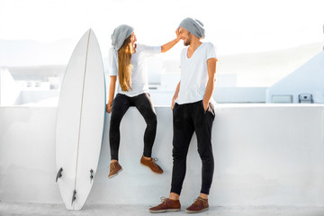 Young couple dressed alike in white t-shirts and hats enjoying summer vacations with surfboard on the roof top in the morning