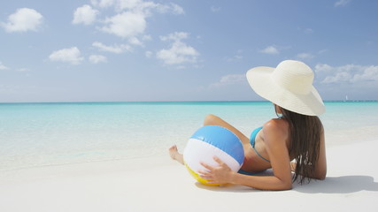 Beach Woman Relaxing Lying In Sand in Paradise on sea shore. Female is wearing blue bikini and sunhat while sunbathing. Beautiful woman holding beach ball on summer vacation holidays. RED EPIC. - Powered by Adobe