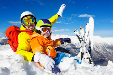 Dad and son skiers sit in snow on mountain top 
