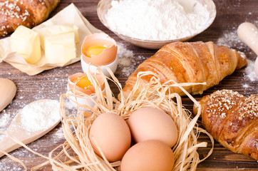 Ingredients for baking croissants - paper, flour, wooden spoon, rolling pin, eggs, egg yolks, butter served on a rustic wooden tray table.
