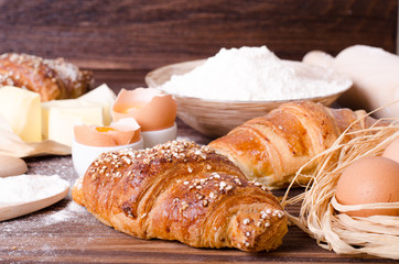 Ingredients for baking croissants - paper, flour, wooden spoon, rolling pin, eggs, egg yolks, butter served on a rustic wooden tray table.