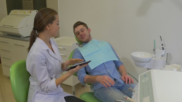 Dentist Clicks a Tablet Shows to a Patient Client is Smiling Pointing on a Tablet Choosing Something Dental Clinic Young Female Doctor Male Client