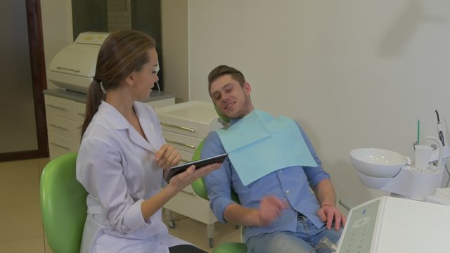 Dentist Scrolls a Tablet Shows to a Patient Client is Talking Pointing on a Tablet Choosing Something Dental Clinic Young Female Doctor Male Client