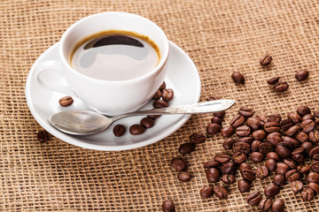 Beautiful white coffee cup and saucer and coffee beans on the background of burlap. Dark background.