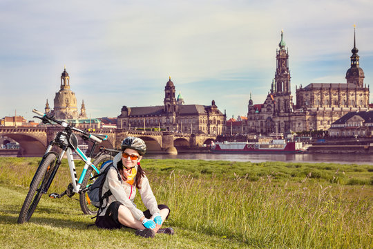 Adult attractive female cyclist sitting on the grass and resting. The happy woman sits at the old city background and looks at the camera. So-called The Balcony of Europe, Dresden, Germany