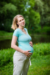 Beautiful smiling young pregnant woman wearing turquoise t-shirt and beige pants with blond hair holding her belly in summer park. Pregnancy and femininity concept. Mother's Day