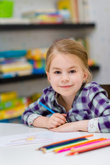 Lovely smiling little girl with blond hair sitting at white table with multicolored pencils and looking at camera