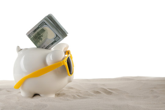 Piggy bank with inserted dollar banknotes and sunglasses on a sand, isolated on white. Holiday money concept