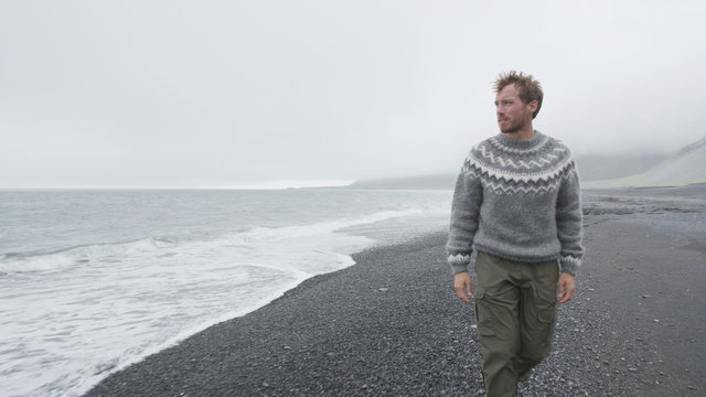 Young man walking on black sand beach by mountain wearing Icelandic sweater on Iceland. Male is in warm clothing during vacation. Man is on walking sea shore.