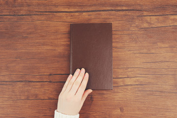 Female hand holding a brown book cover on the brown wooden desk, top view