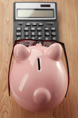 Pink piggy bank with glasses and calculator on wooden background