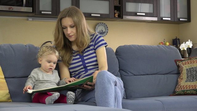 Careful mother showing images in book to her cute little daughter sitting on sofa at home. Toddler child education. Static closeup shot.
