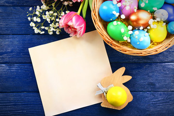 Colorful Easter eggs with flowers and blank greeting card in wicker nest on wooden table, top view