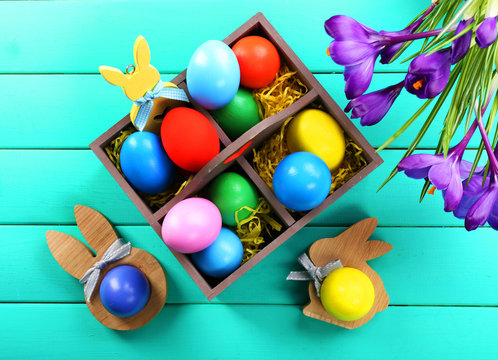 Colorful Easter eggs in box with crocus flowers on wooden table, top view