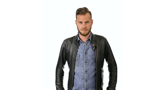 A trendy man wearing a jeans shirt and a black leather jacket, standing in front of a white background looking at camera, feeling calm and laid back.