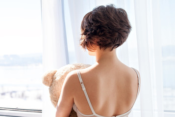 A girl with short hair in a bra back to camera looking out the window and holding a soft toy bear....