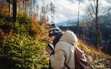 young guy in a warm cap and a jacket with a scarf on the neck, trousers and rubber boots walks alone in the Carpathian mountains with a camera, a tripod and a backpack on his shoulders
