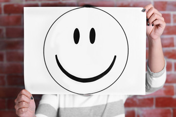 Woman showing a happy emoticon in front of face against a brick wall background
