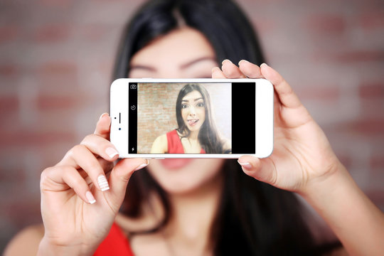 Young attractive girl making photo by her self with mobile phone on brick wall background, close up