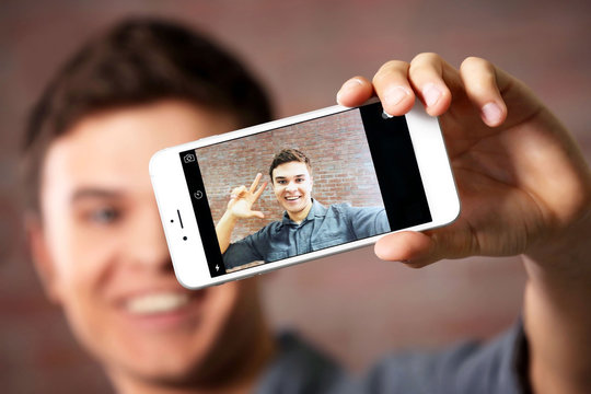Young handsome boy making photo by his self with mobile phone on brick wall background, close up