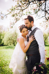 Elegant stylish groom lovely embraces with his happy gorgeous brunette bride on the background of trees in the park. Groom with beard