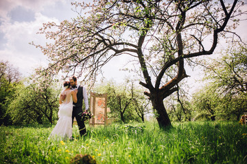 Elegant stylish groom lovely embraces with his happy gorgeous brunette bride on the background of trees in the park. Groom with beard