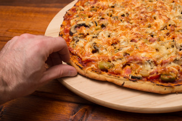The hand reaches for a piece of Italian pizza with ham and mushrooms on a wooden board on natural wooden table