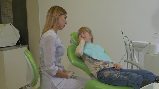 Female Dentist is Talking to Kid Touching Cheek Examining a Teeth of a Patient Kid is Sitting in a Green Chair Dental Clinic Visit to a Stomatologist