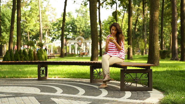 Young beautiful woman holding mobile phone and sitting on bench in summer park