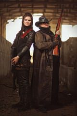 Man with rifle in a leather garment and woman in raider costume with crossbow inside a concrete...