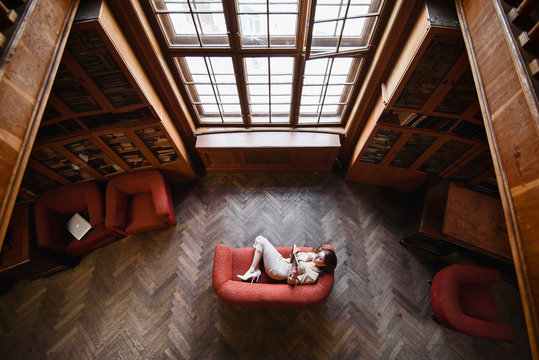 Stylish Young Girl In Beige Suit Reads A Book On The Red Sofa. Top View