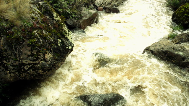 A high-angle view of a turbulent muddy river rushing through a rocky mountain gorge in the Ecuadorian Andes, moss-covered boulders along the banks and churning whitewater.