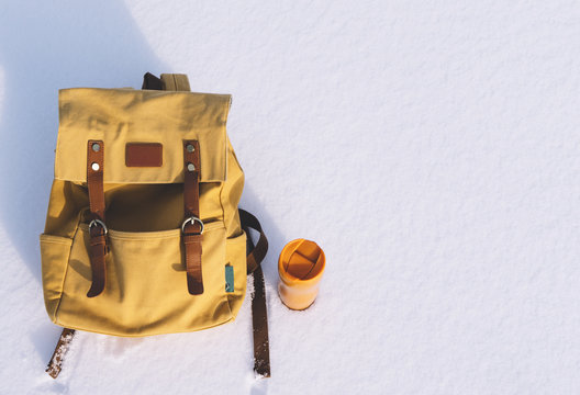 Orange backpack with leather elements and yellow thermos of hot tea or coffee on a background of white pure snow in winter mountains with empty space for text
