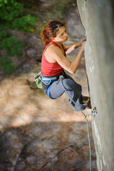 Young beautiful woman climber climbing steep rock with rope, searching for next grip. Top view on focused muscular female athlete. Climbing equipment.