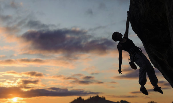 Silhouette of athletic woman rock climber climbing steep rock wall against amazing sunset sky in the mountains on high altitude. Girl is hanging on one hand