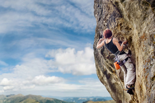Rear view of young female climber climbing with rope and carbines on a big rocky wall against blue sky and mountains. Summer time. Climbing equipment.