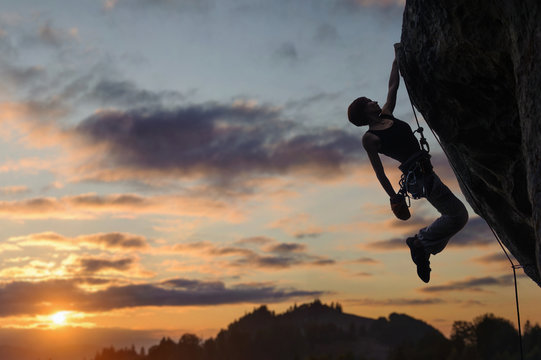 Silhouette of athletic woman rock climber climbing steep rock wall with rope against amazing sunset sky in the mountains. Girl is hanging on one hand and holding hand in magnesium bag.