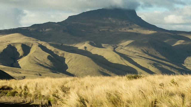 A wide time-lapse of the Corazon volcano in the Ecuadorian Andes, its peak partly shrouded in clouds over a foreground of golden grass swaying in the wind, light and shadow shifting.