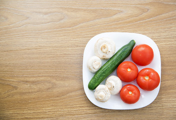 White plate with fresh tomatoes, mushrooms and cucumbers on a wooden table, top view
