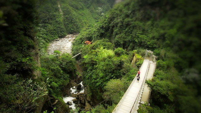 A high-angle view of two hikers crossing a wooden suspension bridge over a deep verdant gorge, a winding river and dense tropical forest below at the Pailon del Diablo near Banos, Ecuador.