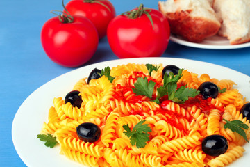 fusilli pasta and olives in the dish near fresh tomato and pieces of bread on a saucer on a wooden blue background