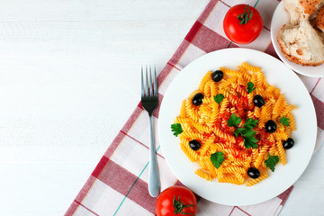 fusilli pasta and olives in a dish near tomatoes and plates with bread on napkin on wooden white background