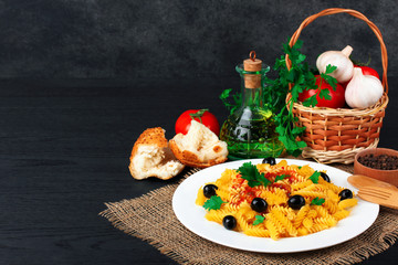 fusilli pasta and olives with parsley and ketchup in the plate near the wicker baskets with tomatoes and garlic with a glass  bottle of sunflower oil on wooden black background