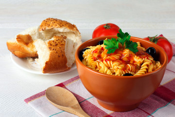 fusilli pasta and olives with parsley and ketchup in a bowl near to the tomatoes and parsley with chunks of fresh bread on a wooden white background