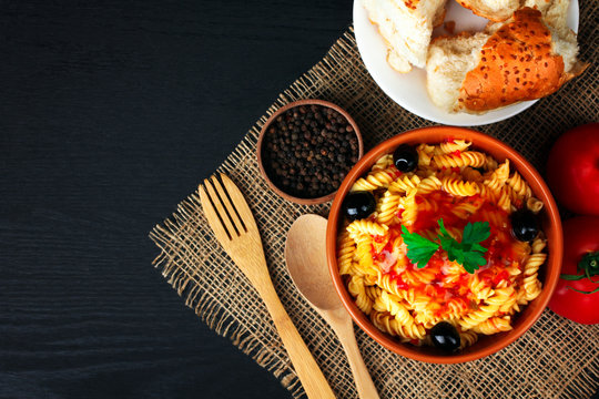 fusilli pasta and olives with parsley and ketchup in a bowl near to the tomatoes and peppers with slices of fresh bread lying on a plate on a wooden black background