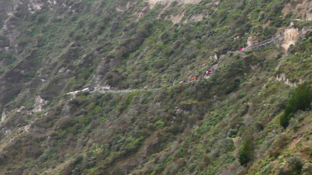 An aerial tracking shot of a winding mountain trail with hikers ascending a rugged green cliffside of the Quilotoa crater in the Ecuadorian Andes, revealing the turquoise lake below.